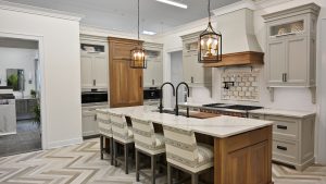 A modern kitchen featuring light gray cabinetry, a central island with a marble countertop, and four upholstered bar stools. Two pendant lights hang over the island, and the backsplash has a subway tile design. Chevron-patterned flooring adds a stylish touch.
