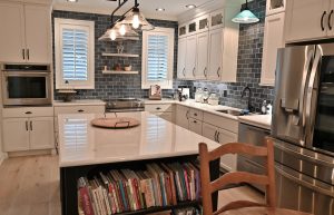 A modern kitchen with white cabinets, a central island with a countertop and built-in bookshelf, and stainless steel appliances. The backsplash is made of dark subway tiles, and two windows with white shutters provide natural light. A pendant light hangs overhead.