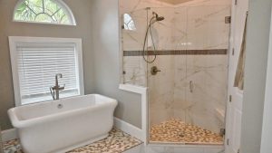 A modern bathroom featuring a freestanding white bathtub with a floor-mounted faucet, positioned near a window with white blinds. Adjacent to the tub is a spacious walk-in shower with glass doors, tiled walls, and a pebble stone floor.