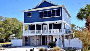 A blue, two-story elevated house with white trim and shutters sits on a neatly landscaped yard. The ground level features an open garage and storage area with brick pillars. A wraparound balcony extends from the second floor, and a staircase leads up to a white gate fence.