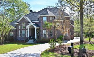 A large, two-story brick house with tall windows and a dark-colored roof. The house is surrounded by green trees and a well-maintained lawn. A concrete driveway leads up to the entrance, and a black mailbox is positioned near the driveway’s edge.