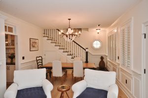 A dining room with a wooden table and six cushioned chairs, two of which have blue cushions. Two white armchairs face the table. A chandelier hangs from the ceiling, and a staircase is in the background. The room has white walls, paneled wainscoting, and a small octagonal window.